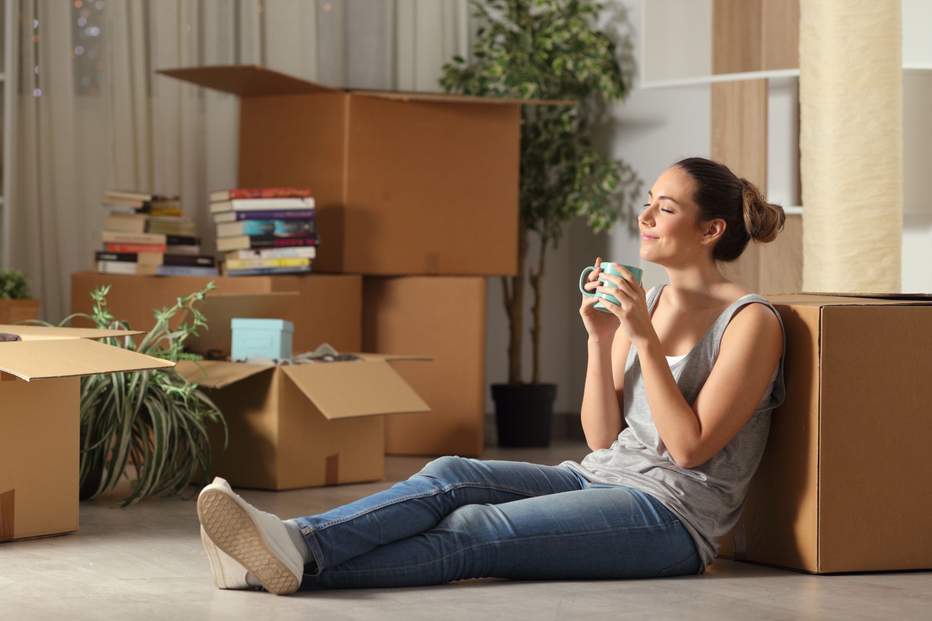 Woman sat by cardboard boxes drinking from cup and smiling